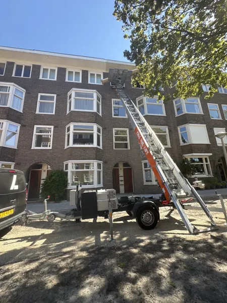 OranjeLift-aanhangerlift achter zwarte bus, ladder tot de dakrand van bakstenen flat. Familiebedrijf dat eerlijk zaken doet tegen een vaste prijs.