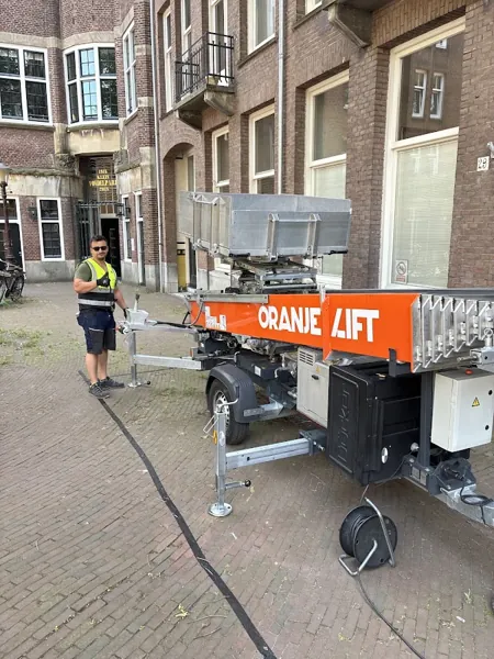 Aanhangerlift van OranjeLift opgesteld op straat bij bakstenen pand, verhuizer erbij. Familiebedrijf dat eerlijk zaken doet tegen een vaste prijs.