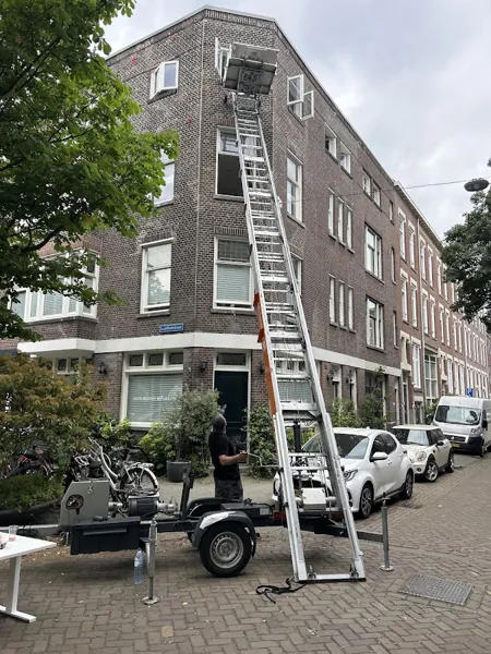 Donker bakstenen hoekpand met aanhangerlift, ladder naar de bovenste etage, bewolkt. Vandaag gebeld, morgen staat OranjeLift al voor je deur.
