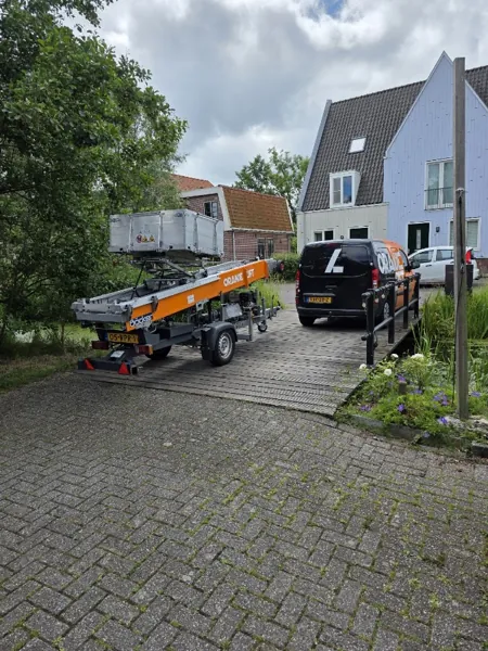 Aanhangerlift en zwarte OranjeLift-bus op houten brug bij houten woning met puntgevel. Piano of Amerikaanse koelkast, wij tillen het veilig omhoog.