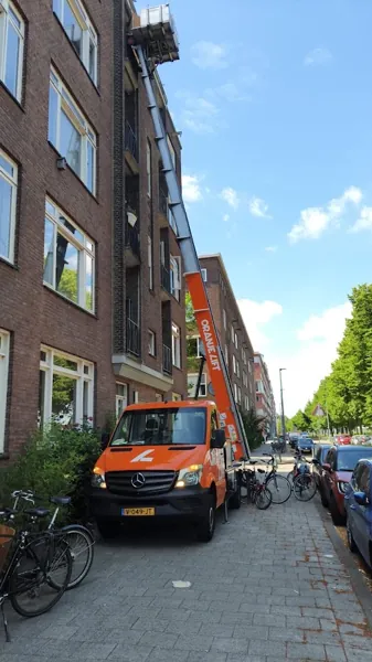 Oranje OranjeLift-vrachtwagen met ladder omhoog langs bakstenen flat met balkons. Onze ladderlift hijst zware kasten zo de hoogte in.