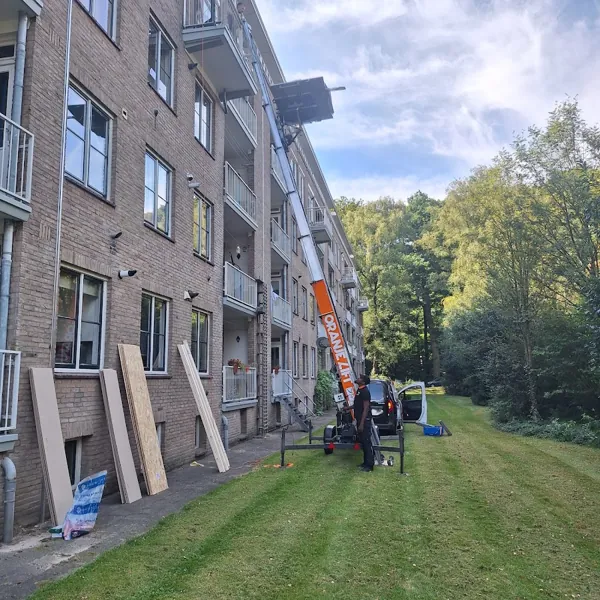 Op het gras naast een bakstenen flat, aanhangerlift met ladder tot bovenste balkon. Bij OranjeLift huur je een verhuislift altijd met operator.