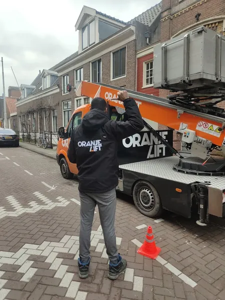 Verhuizer in OranjeLift-jas naast de vrachtwagen met ladderlift in ruststand, bewolkt. Piano of Amerikaanse koelkast, wij tillen het veilig omhoog.