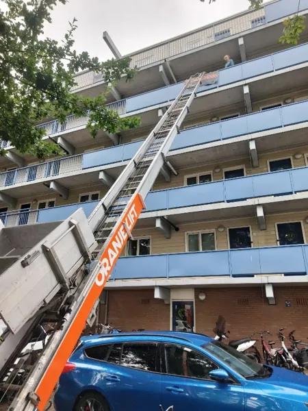 OranjeLift-ladder omhoog naar bovenste galerij van een flat met blauwe balkonhekken. Een appje, en je verhuislift in de regio is geregeld.