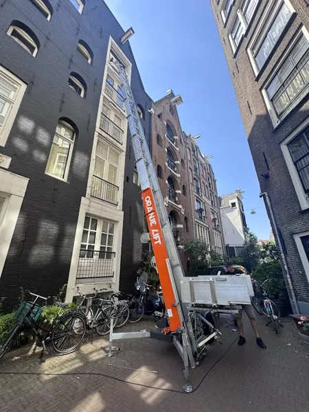 Smalle straat, aanhangerlift met ladder naar bovenste raam van zwart geverfd pand. Vandaag gebeld, morgen staat OranjeLift al voor je deur.