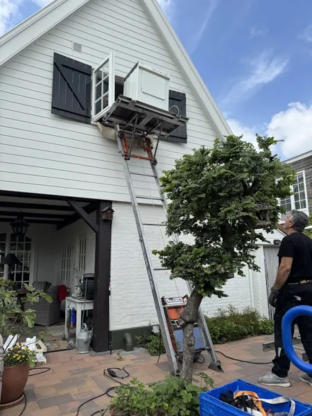 GEDA-lift tegen wit houten huis brengt witgoed naar een open raam met zwarte luiken. Bij OranjeLift huur je een verhuislift altijd met operator.