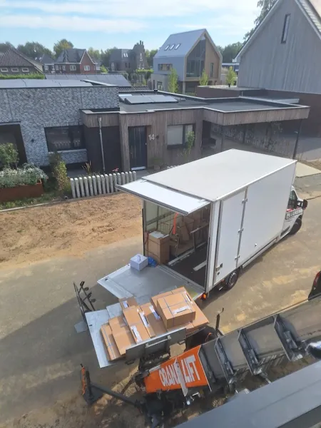 Dozen op het OranjeLift-platform bij een open bakwagen in nieuwbouwwijk, van bovenaf. Voor een smalle straat sturen we de stille aanhangerlift.