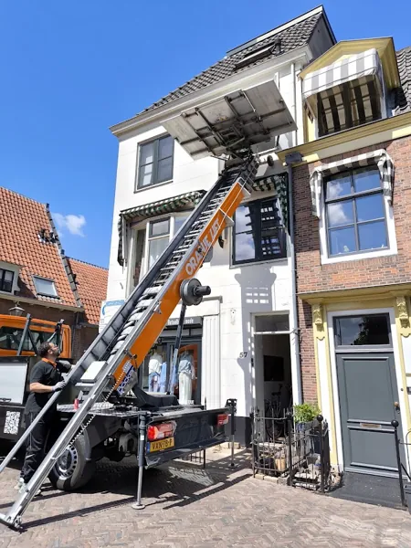 Vrachtwagen met ladderlift omhoog naar de zolder van een wit winkelpand met markiezen. Vandaag gebeld, morgen staat OranjeLift al voor je deur.